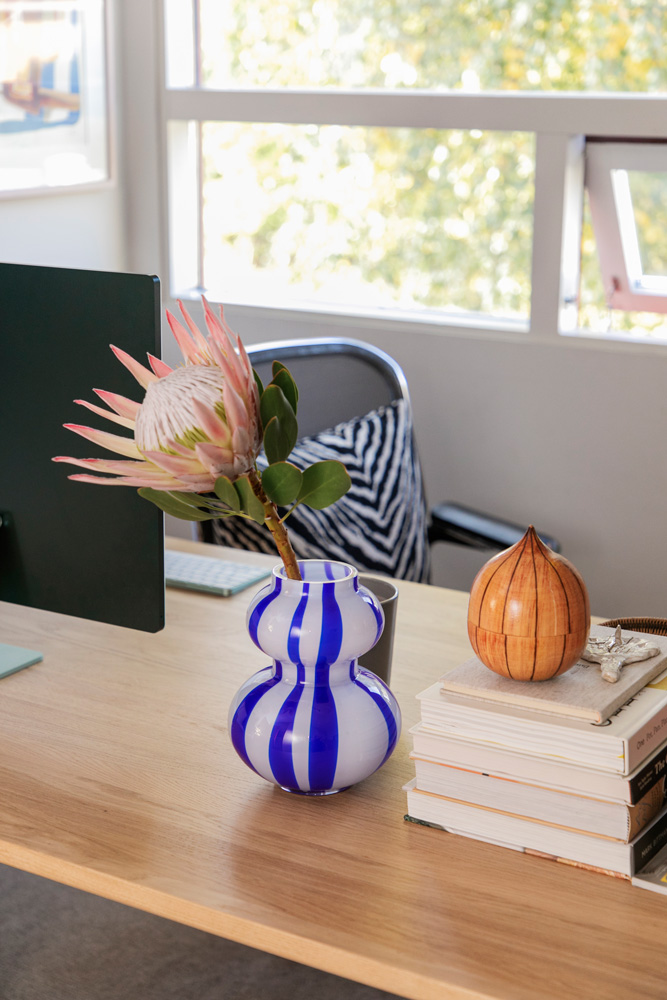 Työpöydällä on asetelma, jossa yhdistyvät luonnon elementit ja moderni muotoilu. Vaalean puisen pöydän pinnalla on sinivalkoraidallinen, kurpitsamainen maljakko, jossa on yksi vaaleanpunainen protea-kukka vihreine lehtineen. Maljakon vieressä on harmaa muki. Pöydällä on myös pino kirjoja, joiden päällä on pieni, oranssi ja raidallinen koriste-esine, joka muistuttaa sipulia tai kurpitsaa. Taustalla näkyy osa tietokonenäyttöä ja harmaa työtuoli, jossa on mustavalkoinen seeprakuvioinen tyyny. Suuri ikkuna tuo luonnonvaloa tilaan, ja sen takana näkyy epäselvästi vihreitä puita ja rakennuksia. Työpöydällä on myös vaaleanvihreä hiirimatto ja mahdollisesti muita työvälineitä, jotka jäävät osittain näkyviin. Asetelma on valoisa ja tyylikäs.