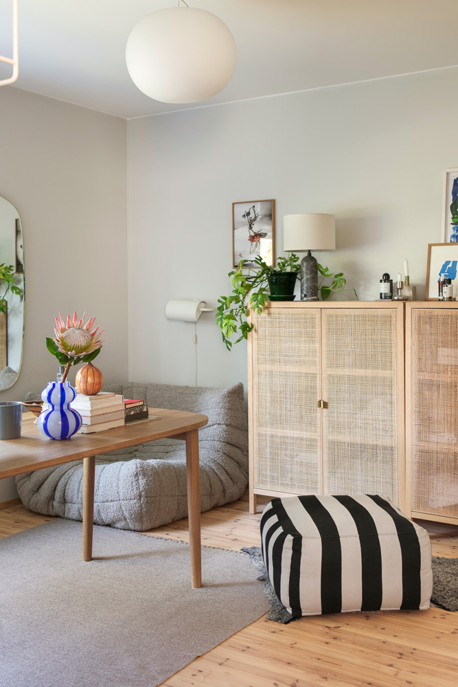 Tässä teksti, johon korjasin pari virhettä: Vaaleassa huoneessa on yhdistetty olohuone- ja ruokailutila. Huoneessa on vaaleanharmaat seinät ja osittain vaalea puulattia, osittain harmaa matto. Ruokailutilassa on vaalea puinen ruokapöytä, jonka päädyssä on harmaa nojatuoli. Pöydällä on sinivalkoinen raidallinen maljakko, jossa on vaaleanpunainen protea-kukka ja pari muuta koristeellista esinettä. Pöydällä on myös muutama kirja ja harmaa muki. Olohuoneen puolella on vaaleanpuinen kaappi rottinkiovilla. Kaapin päällä on harmaa, kivinen jalkalamppu valkoisella lampunvarjostimella, vihreä kasvi vihreässä ruukussa ja muita koriste-esineitä. Kaapin vieressä on mustavalkoraidallinen rahi. Seinällä kaapin yläpuolella on kaksi kehystettyä taulua. Huoneen katossa roikkuu pyöreä, valkoinen paperivalaisin. Peili seinällä heijastaa vihreää kasvia. Huoneessa on rauhallinen ja boheemi tunnelma.