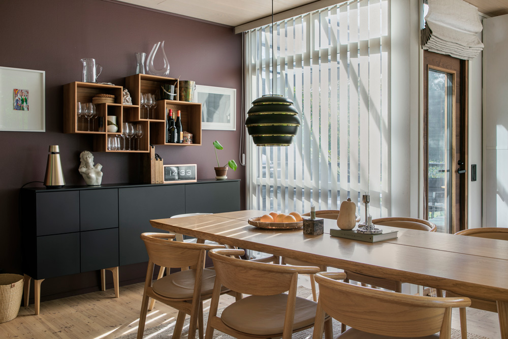 Ruokailutilassa on pitkä, vaalea puinen ruokapöytä, jonka ympärillä on useita vaaleita puutuoleja, joissa on vaaleat pehmusteet. Pöydällä on erilaisia koriste-esineitä, kuten hedelmiä sisältävä kulho, keraamisia astioita ja kynttilänjalka. Pöydän yläpuolella roikkuu moderni, kerroksellinen musta ja kultainen riippuvalaisin. Huoneen seinät ovat tumman purppuranväriset, ja seinää vasten on musta senkki, jonka päällä on kultainen kaiutin, valkoinen pieni patsas ja digitaalinen kello, joka näyttää ajan 12:33. Senkin yläpuolella on epäsymmetrinen, avoin puinen hyllykkö, jossa on erilaisia lasiastioita, keramiikkaa ja viinipulloja. Seinällä on myös kaksi kehystettyä taulua. Huoneessa on suuret ikkunat, joissa on valkoiset pystylamellikaihtimet, ja valo siivilöityy niistä luoden lattialle raitoja. Lattiat ovat vaaleaa parkettia. Huoneessa on moderni ja tyylikäs tunnelma.