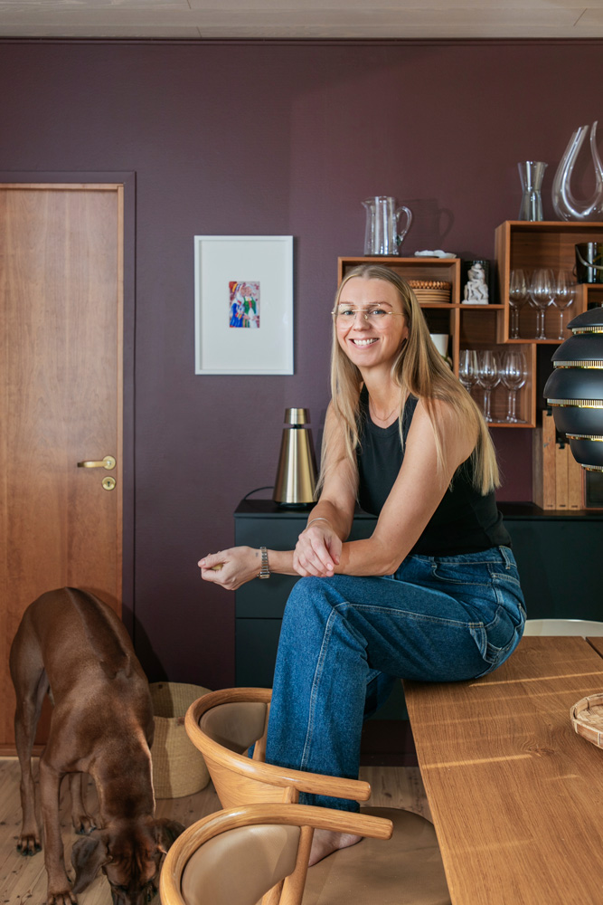Vaaleatukkainen nainen istuu hymyillen puisen pöydän reunalla, jalat vaaleanruskealla tuolilla. Hänellä on yllään musta hihaton toppi, siniset farkut ja vaaleat silmälasit. Hänen takanaan on tumman purppuranvärinen seinä, jossa on valkokehyksinen taulu. Seinää vasten on musta senkki, jonka päällä on kultainen kaiutin ja jonka yläpuolella on avohyllykkö puisilla hyllyillä. Hyllyillä on erilaisia lasiastioita ja koriste-esineitä. Kuvan vasemmassa reunassa näkyy ruskea koira nuuskimassa lattiaa vaalean puuoven vieressä. Huoneessa on lämmin ja kutsuva tunnelma.