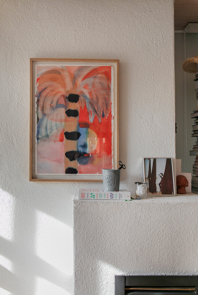 Lähikuvassa näkyy valkoisen, rosoisen seinän päällä oleva matala hylly tai reunus. Seinällä roikkuu vaaleanpuinen kehys, jonka sisällä on abstrakti akvarellimaalaus. Maalauksessa on oransseja, sinisiä ja punaisia sävyjä, ja se esittää mahdollisesti palmua auringonlaskussa. Hyllyllä on useita koriste-esineitä. Vasemmalla on harmaa, kuvioitu metallipurkki, jonka sisällä on tummat sakset. Purkin vieressä on kaksi valkoista kirjaa, joiden selkämyksissä on vihreää ja punaista tekstiä. Oikealla on pieni, neliönmuotoinen valkoinen tuoksukynttilä, jonka vieressä on kaksi pientä kehystettyä kuvaa tai valokuvaa tummilla aiheilla. Aivan oikeassa reunassa näkyy osittain pieni, ruskea saviesine. Seinän alareunassa näkyy musta aukko, joka saattaa olla osa takkaa tai muuta rakenteellista elementtiä. Valo osuu ylhäältä vasemmalta, luoden pehmeitä varjoja esineiden taakse.