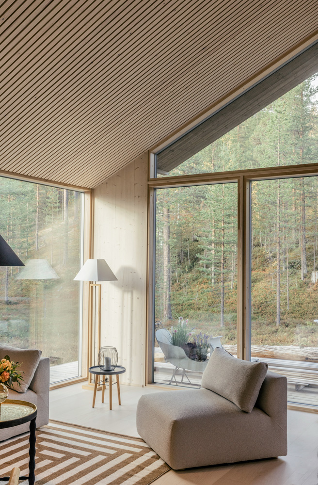 Tilava olohuone, jossa on suuret ikkunat, joista avautuu näkymä metsään. Sisäkatto ja seinät ovat vaaleaa puupaneelia. Huoneessa on vaaleanruskea Hakolan Frame-matto, vaalean beigen värinen Hakolan Puzzle-nojatuoli ja lattiavalaisin. Ikkunoiden kautta näkyy terassi ja metsä.