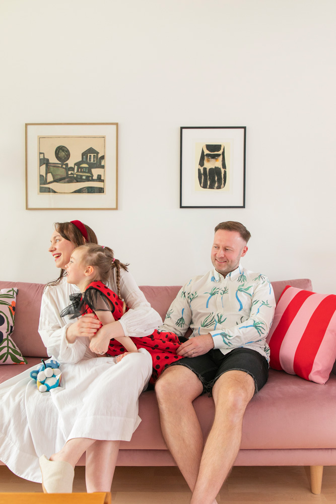 Valokuvassa on perhe, joka istuu vaaleanpunaisella sohvalla. Äiti on pukeutunut valkoiseen mekkoon, ja hän pitelee sylissään lasta, jolla on punainen, pilkullinen mekko. Isä on pukeutunut mustiin shortseihin ja vaaleaan, kuviolliseen paitaan. Olohuoneen seinällä on kaksi kehystettyä taulua. Sohvalla on myös useita tyynyjä.