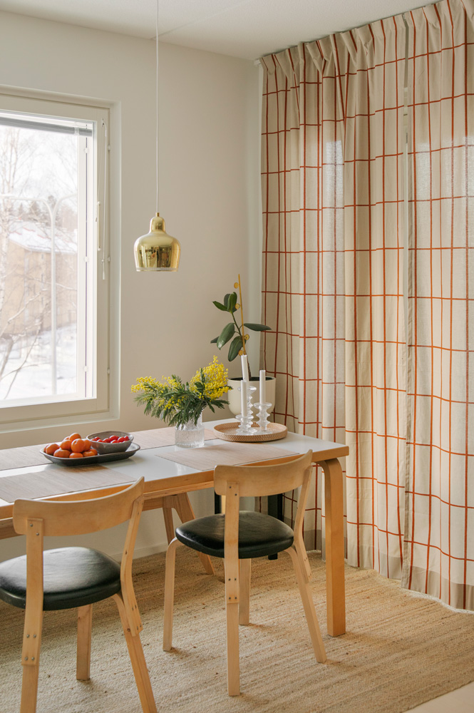 Kuvassa on ruokailutila, jossa on ikkuna vasemmalla ja pitkät, vaaleat, oranssiraitaiset verhot oikealla. Keskellä on vaaleaa puuta oleva ruokapöytä, jonka ääressä on kaksi samanlaista tuolia, joissa on puurunko ja tummat istuinpehmusteet. Pöydän päällä on maljakossa keltaisia kukkia ja kulhossa hedelmiä. Pöydän yläpuolella roikkuu kultainen riippuvalaisin. Lattialla on vaalea juuttimatto.