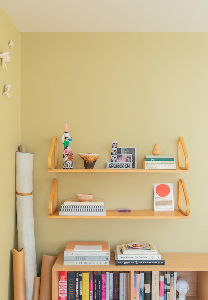Tässä kuvassa on vaaleankeltainen seinä, jonka edessä on hyllykkö. Yläreunassa on kaksi vaaleaa puuhyllyä, joita kannattelevat kaarevat puukannattimet. Hyllyillä on koriste-esineitä, kuten korkea, kirjavasti kuvioitu kynttilä, tummanruskea kulho, valokuvia ja kirjoja. Alhaalla on hyllykkö, jossa on riveittäin kirjoja. Vasemmalla on seinää vasten rullattuja kankaita tai papereita. Vasemmassa yläkulmassa on pieniä, valkoisia, eläimen muotoisia seinäkoristeita.