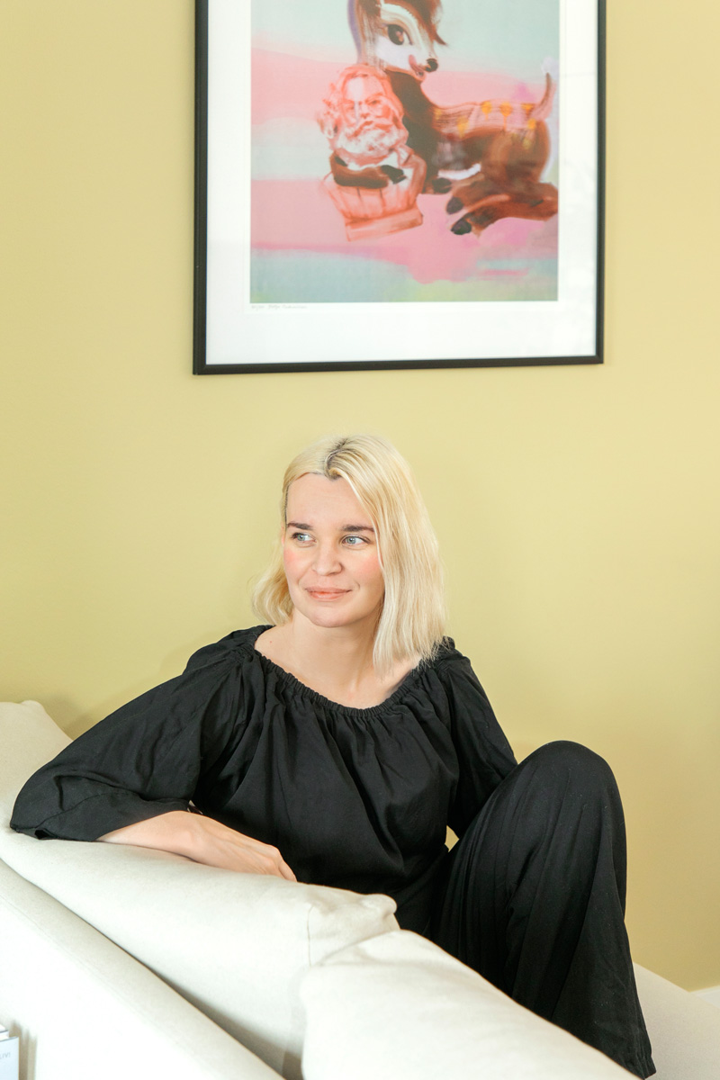 Vaaleahiuksinen nainen, jolla on yllään musta, väljähihainen paita ja mustat housut, istuu valkoisella sohvalla ja katsoo vasemmalle. Hänen takanaan vaaleankeltaisella seinällä roikkuu taulu, jossa on kuva peuranvasasta ja tonttua muistuttavasta hahmosta pastellisävyissä.