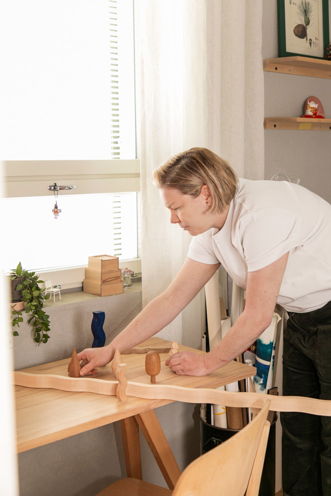Muotoilija Jenni Pehkonen asettelee puisia koriste-esineitä ja veistoksia työpöydälleen valoisassa studiossa. Hänellä on yllään valkoinen t-paita. Pöydällä on abstrakteja puumuotoja ja ikkunasta tulvii pehmeää luonnonvaloa vaaleaan työtilaan.