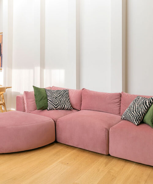 Hakola_friends_Vivian_5_korjattu-pieni Valoisa ja moderni olohuone, jossa pääosassa on vaaleanpunaisella sametilla verhoiltu Hakola Puzzle -moduulisohva. Sohva on koottu L-muotoon, ja siinä on suuri, kaareva avopäätyosa sekä vihreitä ja seeprakuvioisia koristetyynyjä. Lattiana on vaalea parketti. Taustalla on puinen ruokailuryhmä, seeprakuvioiset tuolit, maljakollinen vaaleanpunaisia tulppaaneja ja seinällä abstrakti taideteos. Huone kylpee pehmeässä luonnonvalossa.