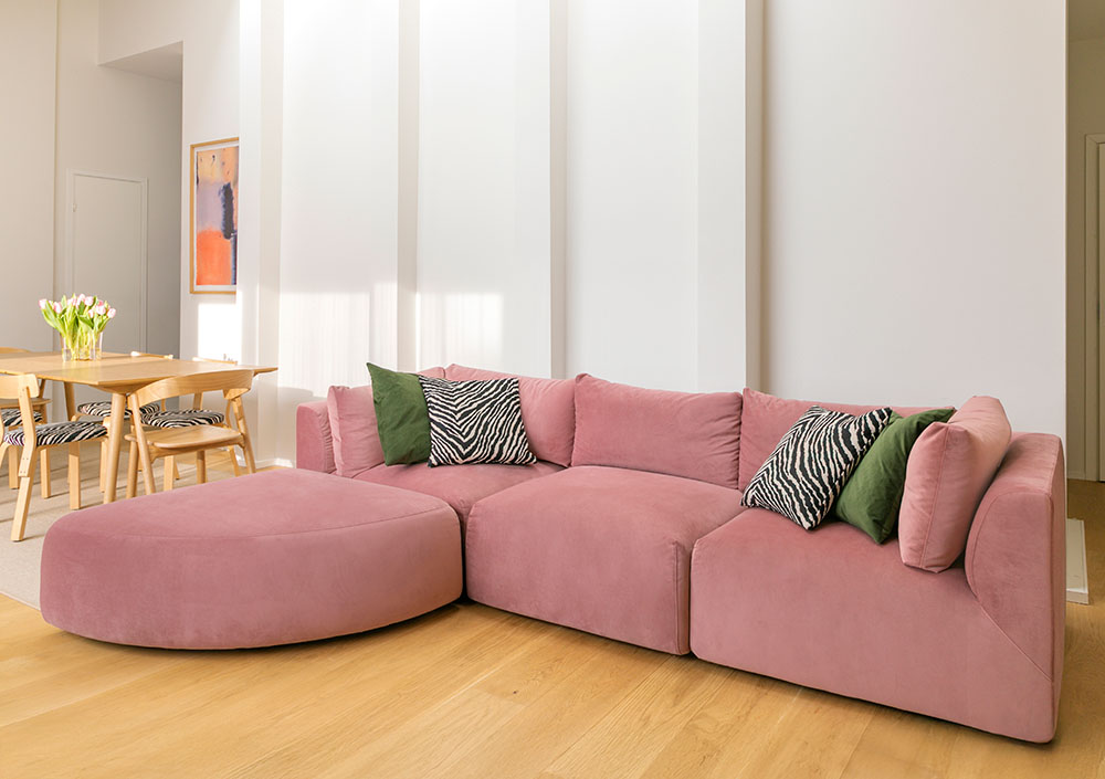 Valoisa ja moderni olohuone, jossa pääosassa on vaaleanpunaisella sametilla verhoiltu Hakola Puzzle -moduulisohva. Sohva on koottu L-muotoon, ja siinä on suuri, kaareva avopäätyosa sekä vihreitä ja seeprakuvioisia koristetyynyjä. Lattiana on vaalea parketti. Taustalla on puinen ruokailuryhmä, seeprakuvioiset tuolit, maljakollinen vaaleanpunaisia tulppaaneja ja seinällä abstrakti taideteos. Huone kylpee pehmeässä luonnonvalossa.