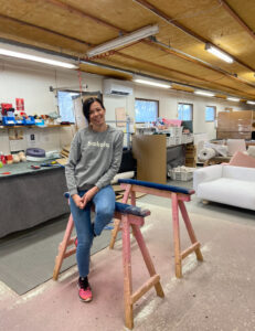 Hymyilevä nainen, Hakolan luova johtaja Annaleena Hakola, istuu rennosti vaaleanpunaisen puupukin päällä Hakolan huonekalutehtaan verhoomossa. Hänellä on yllään harmaa collegepaita, jossa lukee "hakola", sekä siniset farkut ja punaiset lenkkarit. Taustalla näkyy työtaso työkaluineen, kankaita, pehmusteita sekä valmiita huonekalujen osia, kuten valkoinen sohvatyyny. Tilan katossa on loisteputkivalaisimet ja eristevillaa.