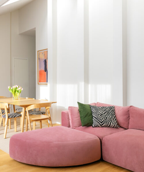 Lähikuva modernista olohuoneesta, jonka keskipisteenä on vaaleanpunaisella sametilla verhoiltu Hakola Puzzle -moduulisohva. Sohvaan on yhdistetty sektorin muotoinen Puzzle-kaarirahi, ja kokonaisuutta täydentävät vihreä ja seeprakuvioinen koristetyyny. Taustalla näkyy ruokailuryhmä, jossa on puiset tuolit, seeprakuvioiset istuimet ja vaaleanpunaisia tulppaaneja maljakossa. Seinällä on abstrakti taideteos. Huone kylpee luonnonvalossa, ja vaalea parketti tuo lämpöä tilaan.