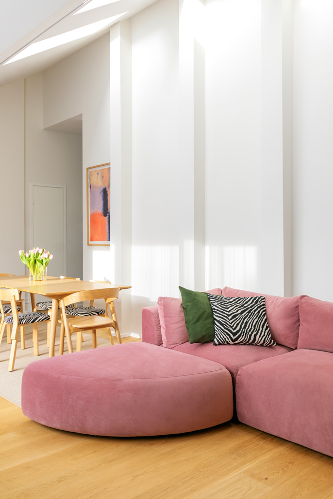 hakola-vivian-2 Lähikuva modernista olohuoneesta, jonka keskipisteenä on vaaleanpunaisella sametilla verhoiltu Hakola Puzzle -moduulisohva. Sohvaan on yhdistetty sektorin muotoinen Puzzle-kaarirahi, ja kokonaisuutta täydentävät vihreä ja seeprakuvioinen koristetyyny. Taustalla näkyy ruokailuryhmä, jossa on puiset tuolit, seeprakuvioiset istuimet ja vaaleanpunaisia tulppaaneja maljakossa. Seinällä on abstrakti taideteos. Huone kylpee luonnonvalossa, ja vaalea parketti tuo lämpöä tilaan.