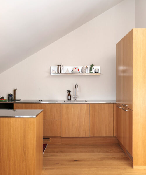 hakola-vivian-3 Yleiskuva vaaleapuisesta keittiöstä, jonka takaseinällä on tasot ja allas, sekä oikealla puolella korkea umpinainen kaapisto. Allasta vastapäätä, kuvan vasemmassa reunassa, näkyy osa keittiösaarekkeesta tai niemekkeestä, jossa on myös ruostumattomasta teräksestä valmistettu työtaso. Takaseinän työtasolla, hanan vieressä, on tumma pullo, ja seinällä on avohylly, jossa on esillä keraamisia astioita, pieni vihreä esine ja kynttilä. Koko tila kylpee luonnonvalossa, ja lattiana on vaalea parketti.