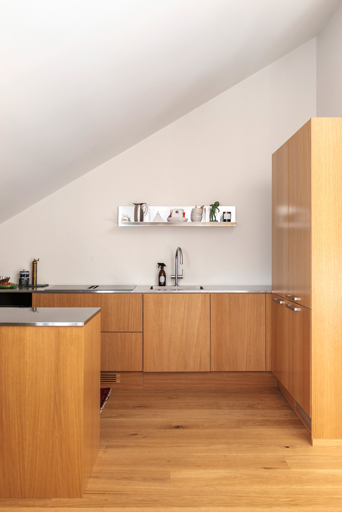 hakola-vivian-3 Yleiskuva vaaleapuisesta keittiöstä, jonka takaseinällä on tasot ja allas, sekä oikealla puolella korkea umpinainen kaapisto. Allasta vastapäätä, kuvan vasemmassa reunassa, näkyy osa keittiösaarekkeesta tai niemekkeestä, jossa on myös ruostumattomasta teräksestä valmistettu työtaso. Takaseinän työtasolla, hanan vieressä, on tumma pullo, ja seinällä on avohylly, jossa on esillä keraamisia astioita, pieni vihreä esine ja kynttilä. Koko tila kylpee luonnonvalossa, ja lattiana on vaalea parketti.