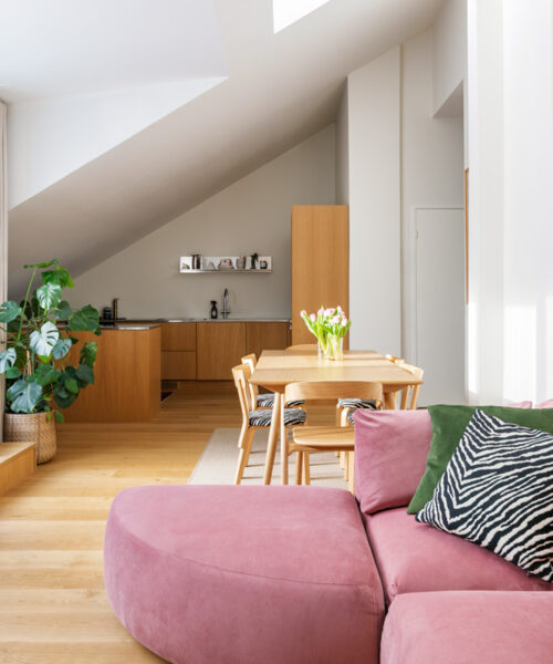 hakola-vivian-4 Yleiskuva valoisasta, loft-tyylisestä asunnosta, jossa on avoin olohuone, ruokailutila ja keittiö. Etualalla on vaaleanpunaisella sametilla verhoiltu Hakola Puzzle -moduulisohva, jonka L-muoto on muodostettu liittämällä sohvaan sektorin muotoinen Puzzle-kaarirahi. Sohvaa koristavat vihreä ja seeprakuvioinen koristetyyny. Olohuoneesta siirrytään ruokailutilaan, jossa on puinen ruokapöytä ja tuoleja sekä vaaleanpunaisia tulppaaneja maljakossa. Taustalla on integroiduilla puukaapeilla varustettu keittiö, joka sijaitsee vinon katon alla. Suuret ikkunat ja kattoikkuna tuovat tilaan runsaasti luonnonvaloa, joka luo pehmeitä varjoja. Vaalea puulattia yhtenäistää tilat.